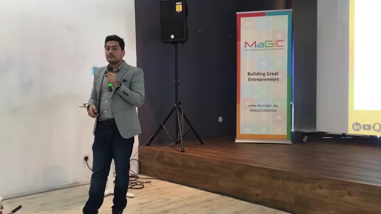 Syed Irfan Ajmal speaking with a handheld microphone beside a MaGIC roll-up banner reading Building Great Entrepreneurs, at MaGIC in Cyberjaya